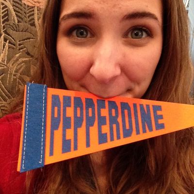 Young woman holding an orange and blue 'Pepperdine' pennant in front of her face, smiling with wide eyes
