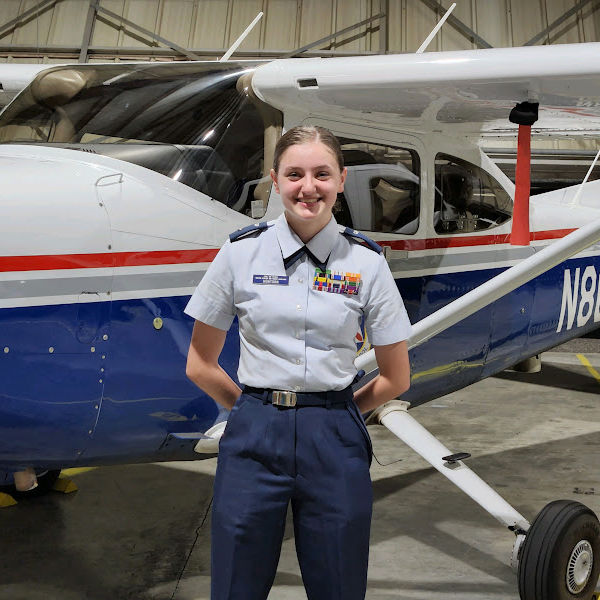 Young woman in military-style uniform standing in front of a small aircraft inside a hangar