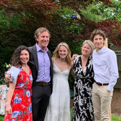 Group of five people smiling together outdoors in semi-formal attire, standing in front of a tree-lined background