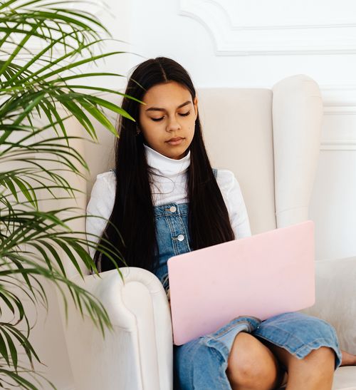 Young girl sitting in a white armchair using a pink laptop, partially hidden behind a leafy green plant