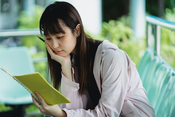 Thoughtful young woman sitting outdoors, reading a yellow notebook and holding a pencil to her cheek