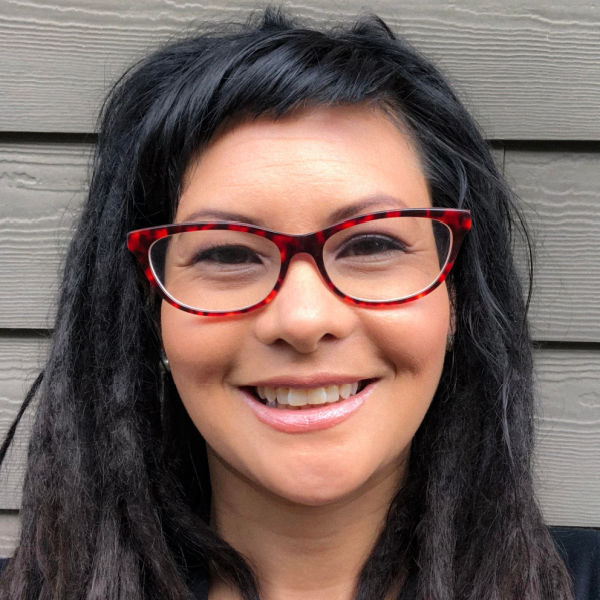 Woman with red glasses and dark dreadlocks smiling in front of gray wood paneling